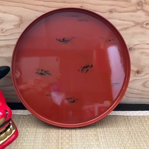 以下が含まれることがあります： 赤色の漆塗りの大きな丸いお盆。黒いアクセントがあります。光沢のある仕上がりで、縁が少し盛り上がっています。食べ物を提供したり、物を飾ったりするのに使われる可能性が高いです。伝統的な日本のデザインです。