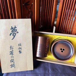 May include: A wooden box with a copper tea set inside. The box is labeled with Japanese characters and the words "tea set". The tea set includes a copper teapot and a copper cup.