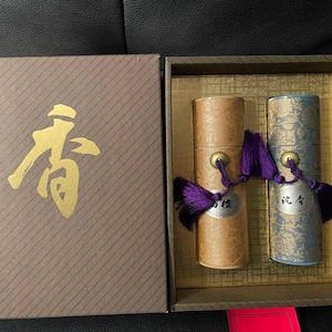以下が含まれることがあります： 蓋に金色の漢字が描かれた茶色の箱で、開くと2つの円筒形の容器が現れます。一つはタン色、もう一つは金色の花柄の青色です。どちらにも紫色のタッセルが付いています。