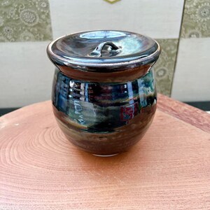 以下が含まれることがあります： 蓋付きの小さな手作りの陶器の壺。壺は、青、緑、茶色の虹色の色合いを持つ、暗く光沢のある仕上がりです。蓋には小さなハンドルが付いています。壺は木製の表面に置かれています。