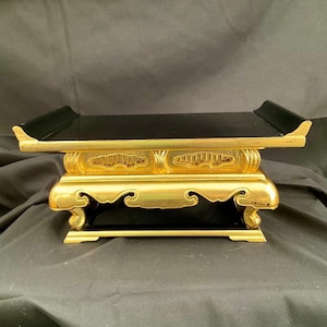 May include: A gold-colored, rectangular decorative stand with a black top and base. The stand features ornate carvings and a curved design on the legs and sides. The top has raised edges.