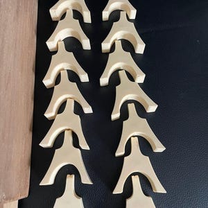 以下が含まれることがあります： 2列に並べられた14個の明るい色のアーチ型木製コンポーネント。各ピースは湾曲した上部と2本の広がった脚が特徴で、滑らかなマット仕上げと一貫した明るいベージュの木目が施されています。