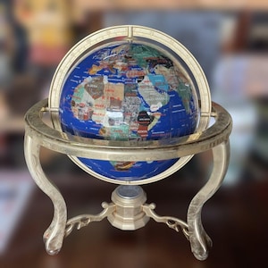 May include: A decorative globe with a blue lapis lazuli sphere, showcasing continents in various stone inlays. The globe is held by a silver-toned metal stand with curved legs and a circular frame, creating an elegant display piece.