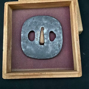 Peut inclure: Une garde d'épée en fer gris foncé patiné, appelée tsuba, présentant une ouverture centrale effilée et deux découpes latérales. La surface métallique est texturée et vieillie. L'objet est présenté dans une boîte en bois à doublure rouge.