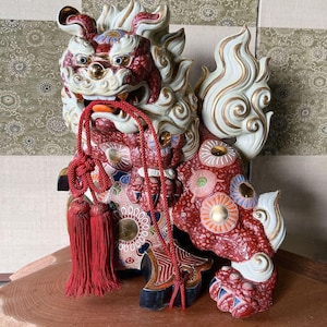 May include: A detailed ceramic guardian lion statue, featuring a red, white, and gold color scheme. The statue has intricate floral and geometric patterns, with a red tassel necklace. The lion is posed on a wooden surface.
