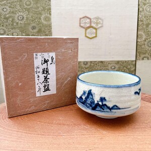 以下が含まれることがあります： 山岳風景のデザインが施された白と青のセラミックティーカップ。カップは日本語のテキストが入った茶色の箱の隣にあります。カップには青い縁があり、木製の表面に置かれています。
