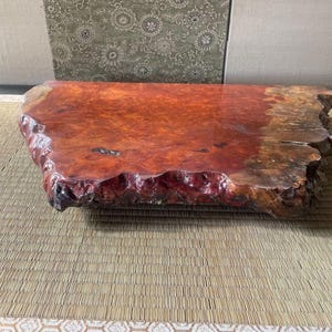 May include: A large, natural wood slab with a reddish-brown color and a rough, irregular edge. The slab is sitting on a woven mat.