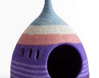 May include: A colorful, felted pet bed in a whimsical, pod-like shape. The bed features horizontal stripes in shades of blue, white, pink, and purple. It has a large circular opening and a curled top. The base is a deep burgundy.