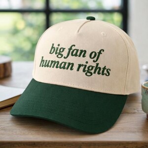 May include: A baseball cap with a cream-coloured crown and a dark green brim. The text "big fan of human rights" is printed in dark green on the front of the cap. The cap is on a wooden surface.