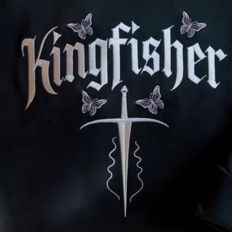 May include: Black garment with white embroidered design. The design features the word "Kingfisher" in a gothic font, a sword, and four butterflies. The embroidery is detailed and the overall aesthetic is elegant.