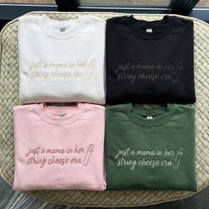 Puede incluir: Cuatro sudaderas de cuello redondo en blanco, negro, rosa y verde. Cada sudadera tiene el texto bordado "just a mama in her string cheese era" con un gráfico de queso en hebra. Las sudaderas están dobladas y expuestas sobre una superficie tejida.