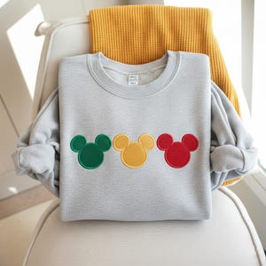 Puede incluir: Una sudadera gris claro con tres dise&ntilde;os bordados de la cabeza de Mickey Mouse en verde, amarillo y rojo. La sudadera est&aacute; doblada sobre una silla con una manta amarilla en el fondo.