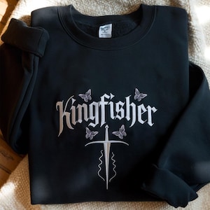 May include: Black sweatshirt with the word "Kingfisher" in white script, accompanied by butterfly and sword designs. The sweatshirt is folded, showcasing the embroidered design on the front. The garment is made of a soft material.