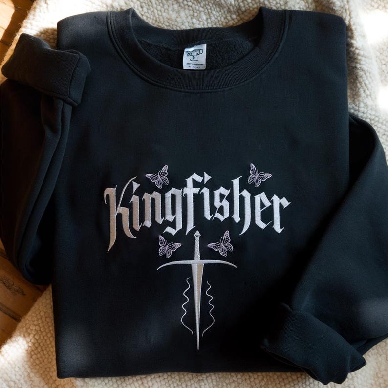 May include: Black sweatshirt with the word "Kingfisher" in white script, accompanied by butterfly and sword designs. The sweatshirt is folded, showcasing the embroidered design on the front. The garment is made of a soft material.