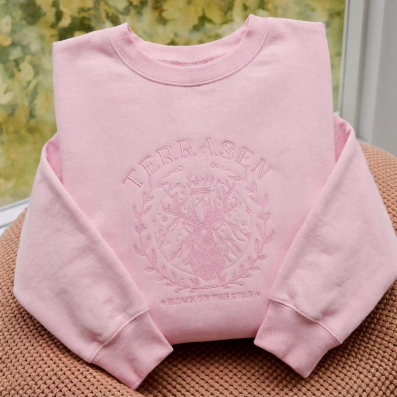 May include: A light pink sweatshirt with the word "TERRASEN" embroidered in a circular design with a deer and the words "HOME OF THE STAG". The sweatshirt is folded on a brown surface.