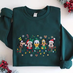 Peut inclure: Un sweat-shirt à manches longues vert foncé avec une broderie représentant cinq personnages de dessin animé portant des accessoires d'hiver. La broderie inclut Dingo, Minnie, Donald, Pluto et Mickey, agrémentée de guirlandes lumineuses.