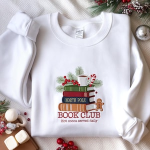 May include: White sweatshirt with an embroidered design of a book stack, a cocoa cup, and a candy cane. The words "North Pole Book Club" and "Hot cocoa served daily" are embroidered on the sweatshirt. Festive winter apparel.