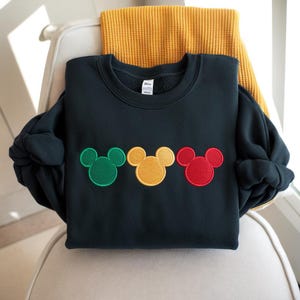 Puede incluir: Una sudadera azul marino oscuro con tres apliques de cabeza de Mickey Mouse en verde, amarillo y rojo. La sudadera est&aacute; doblada sobre una silla con una bufanda amarilla en el fondo.