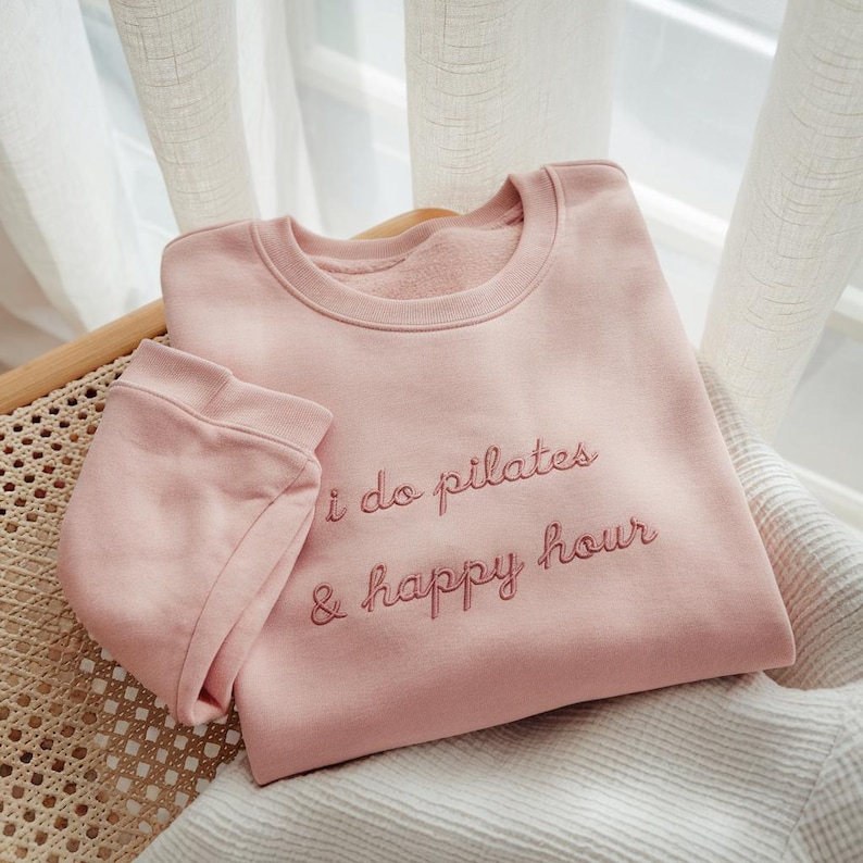 May include: A light pink sweatshirt with the embroidered text "i do pilates & happy hour." The sweatshirt is folded and placed on a woven chair. The sleeves are folded over the body of the sweatshirt.