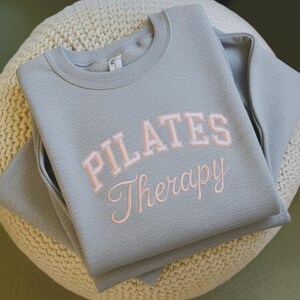 May include: A light gray sweatshirt with the words "PILATES Therapy" in a pink, collegiate-style font. The sweatshirt is folded neatly on a cream-colored, textured surface. The crew neck sweatshirt has long sleeves.
