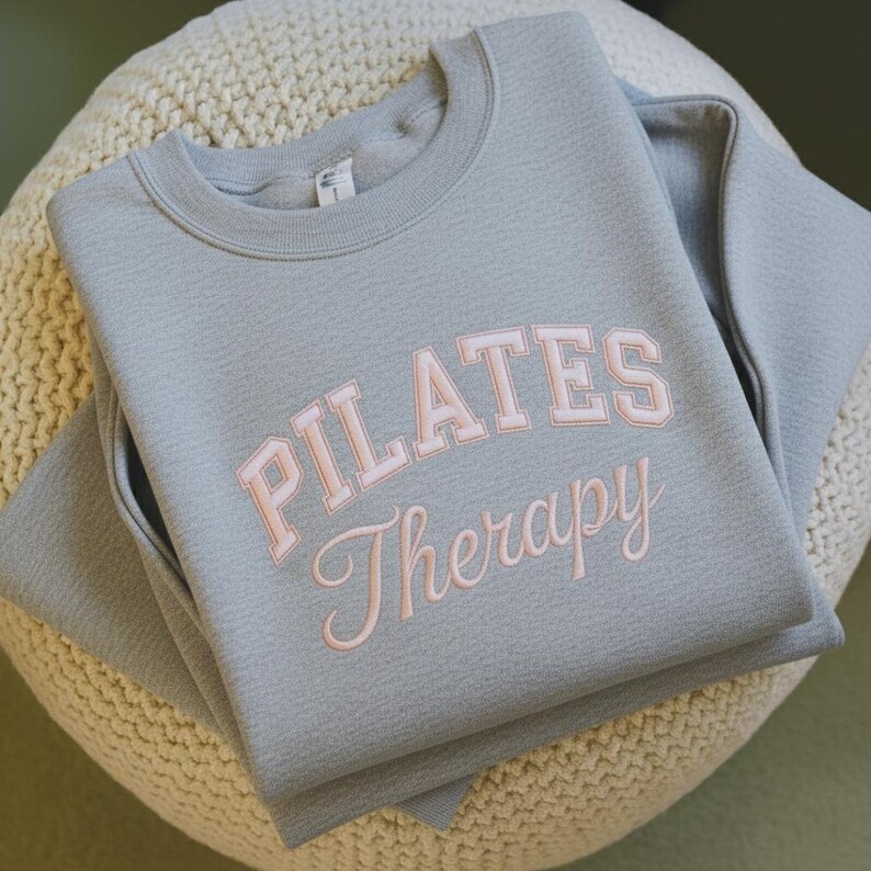May include: A light gray sweatshirt with the words "PILATES Therapy" in a pink, collegiate-style font. The sweatshirt is folded neatly on a cream-colored, textured surface. The crew neck sweatshirt has long sleeves.