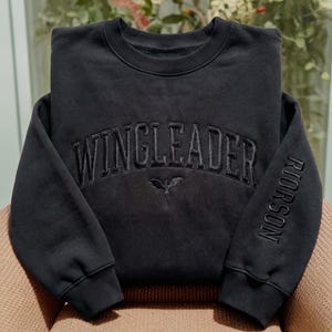 May include: A black crewneck sweatshirt featuring the embroidered text WINGLEADER in an arched font across the chest, with a small dragon icon centred below. The word RIORSON is embroidered vertically along the outer side of the left sleeve.