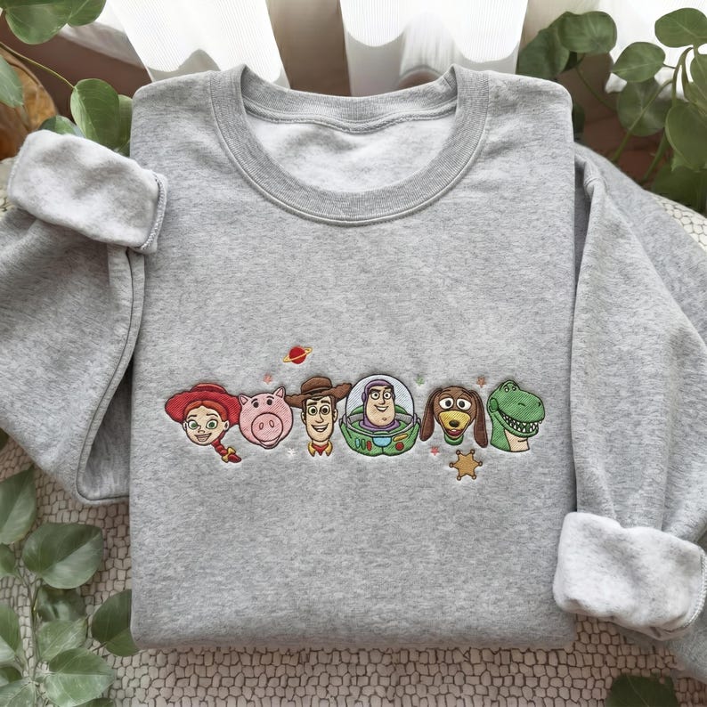 May include: A light gray crewneck sweatshirt featuring an embroidered design of six animated characters including a cowgirl, a pig, a cowboy, a space ranger, a dachshund, and a green dinosaur, accented with a small planet and a sheriff star.