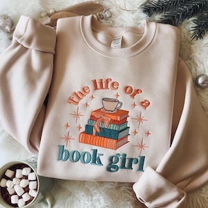 May include: A light pink sweatshirt with the embroidered phrase "the life of a book girl." The design includes a stack of books, a coffee cup, and starburst accents in orange and teal. The sleeves are cuffed.