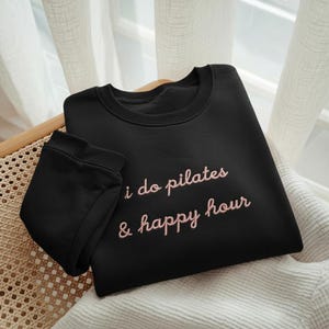 May include: Black sweatshirt with the embroidered text "i do pilates & happy hour" in a pink script. The sweatshirt is folded and placed on a woven chair, with a white curtain in the background.
