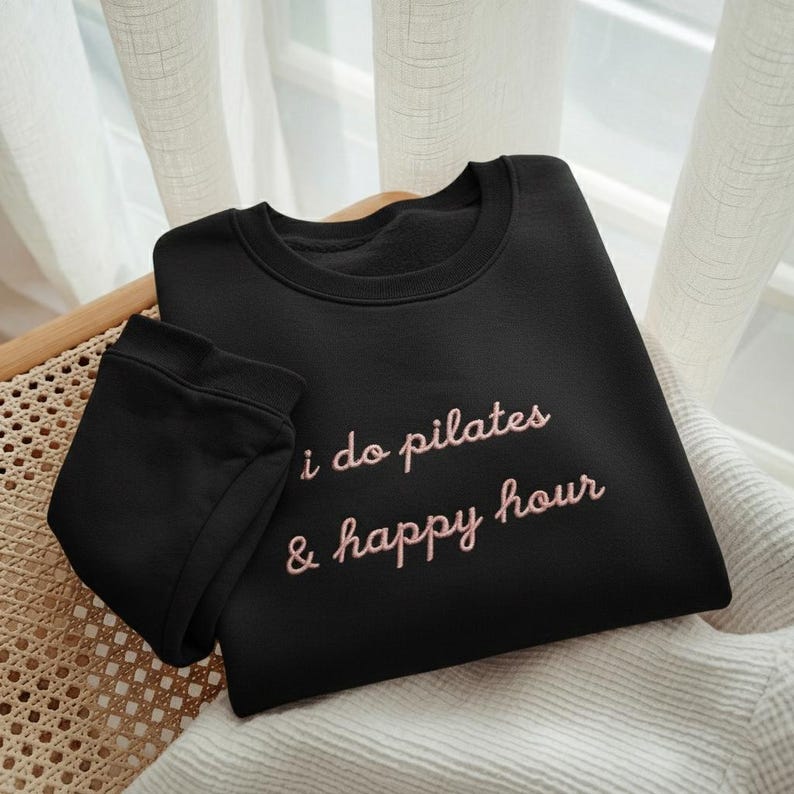 May include: Black sweatshirt with the embroidered text "i do pilates & happy hour" in a pink script. The sweatshirt is folded and placed on a woven chair, with a white curtain in the background.