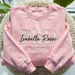 May include: A light pink sweatshirt with the embroidered text "PhD Isabella Rossi Est. 2026". The sweatshirt has a crew neck and long sleeves. A small graduation cap and the word "Education" are embroidered on the left sleeve.