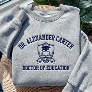 May include: A heather gray crewneck sweatshirt featuring dark navy blue embroidery. The design includes the text DR. ALEXANDER CARTER, a central shield emblem with a graduation cap and an open book, the text EST. 2026, and DOCTOR OF EDUCATION.