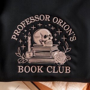 May include: Black sweatshirt with a rose gold embroidered design. The design features a skull atop a stack of books, a lit candle, and the words "Professor Orion's Book Club" in an arched layout. A floral rose is also included in the design.