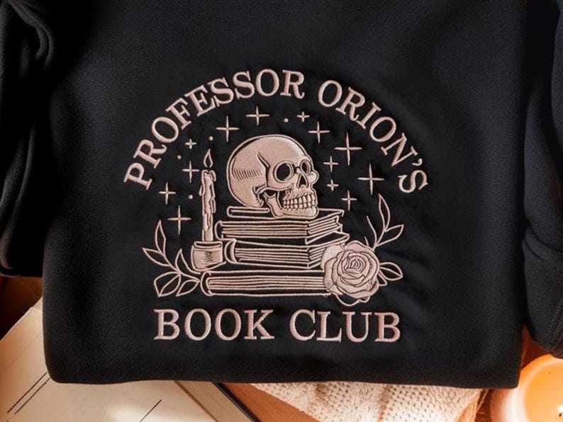 May include: Black sweatshirt with a rose gold embroidered design. The design features a skull atop a stack of books, a lit candle, and the words "Professor Orion's Book Club" in an arched layout. A floral rose is also included in the design.