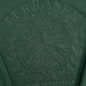 May include: Dark green sweatshirt with an embroidered design. The design features the word "TERRASEN" above a circular emblem with a stag, mountains, and the text "HOME OF THE STAG". The emblem is surrounded by a laurel wreath.
