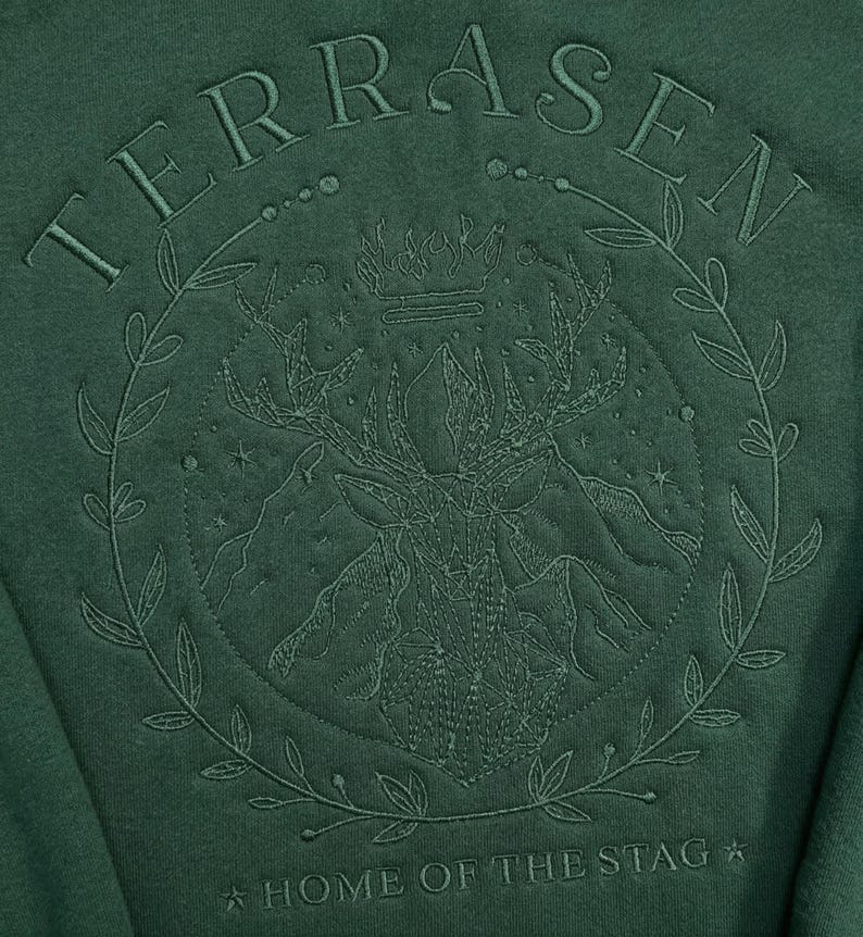 May include: Dark green sweatshirt with an embroidered design. The design features the word "TERRASEN" above a circular emblem with a stag, mountains, and the text "HOME OF THE STAG". The emblem is surrounded by a laurel wreath.