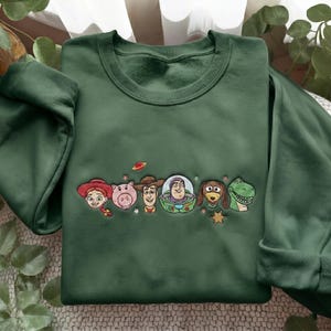 May include: A forest green crewneck sweatshirt featuring an embroidered design of six animated characters from the Toy Story franchise, including Jessie, Hamm, Woody, Buzz Lightyear, Slinky Dog, and Rex, accented with small stars and a planet icon.