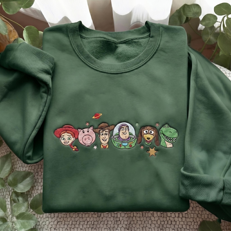 May include: A forest green crewneck sweatshirt featuring an embroidered design of six animated characters from the Toy Story franchise, including Jessie, Hamm, Woody, Buzz Lightyear, Slinky Dog, and Rex, accented with small stars and a planet icon.