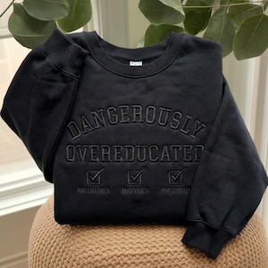 May include: A black crewneck sweatshirt featuring tonal black embroidery. The text reads DANGEROUSLY OVEREDUCATED in an arched font, followed by three checkbox icons with checkmarks and the labels BACHELOR'S, MASTER'S, and DOCTORATE below.