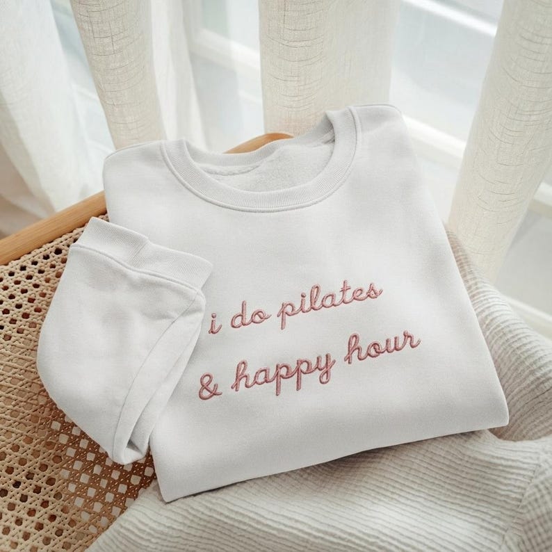 May include: White crewneck sweatshirt with the embroidered text "i do pilates & happy hour" in a pink script. The sweatshirt is folded on a woven chair with a neutral-colored blanket and a sheer curtain in the background.