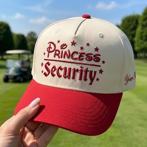 Może przedstawiać: Kremowa czapka z daszkiem z czerwonym daszkiem i czerwonym guzikiem na górze. Z przodu znajduje się wyhaftowany czerwony napis "Princess Security" otoczony małymi czerwonymi gwiazdkami. Z boku czapki wyhaftowano napis "Your Name".