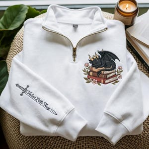 May include: A white quarter-zip sweatshirt featuring an embroidered design on the chest of a black dragon with gold horns sleeping on a stack of books surrounded by flowers. The sleeve features an embroidered sword with the text Violent Little Thing.