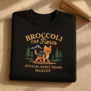Könnte beinhalten: Schwarzer Pullover mit einem gestickten Design eines orangefarbenen Katers mit einem blauen Rucksack, der in einem Wald spazieren geht. Der Text "Broccoli the Fierce" befindet sich über dem Kater und "Official Quest Squad Mascot" unter dem Kater.