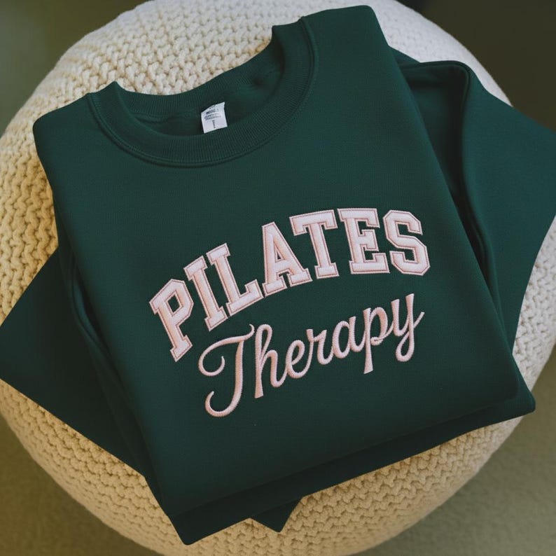 May include: A dark green sweatshirt with the words "PILATES Therapy" in a light pink, collegiate-style font. The sweatshirt is folded neatly, showcasing the design prominently. The garment appears to be made of a soft material, suitable for casual wear.