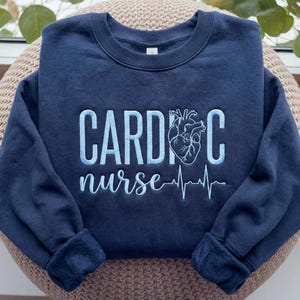 Op de afbeelding: Marineblauw sweatshirt met lichtblauwe borduursels van "CARDIAC nurse", een hartafbeelding en een hartslaglijn. De sweatshirt heeft een ronde hals en lange mouwen.