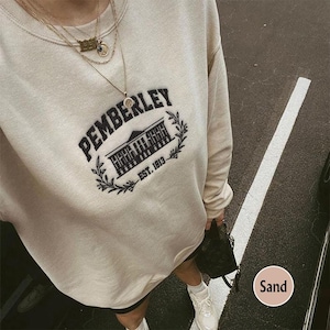 May include: A beige sweatshirt with a black embroidered design that says "Pemberley Est. 1813". The design features a building with a pointed roof and a small circle with a design inside.