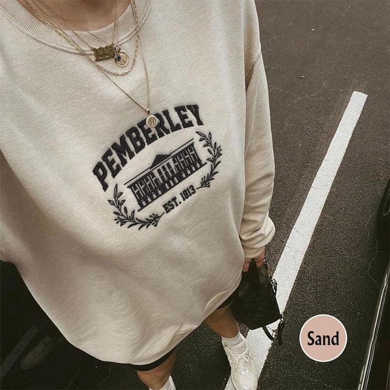May include: A beige sweatshirt with a black embroidered design that says "Pemberley Est. 1813". The design features a building with a pointed roof and a small circle with a design inside.