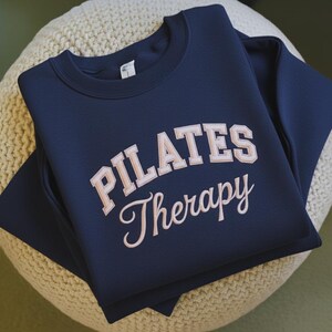 May include: A navy blue sweatshirt with the words "PILATES Therapy" in a light pink, collegiate-style font. The sweatshirt is folded neatly on a beige, textured surface, suggesting a focus on fitness and wellness.