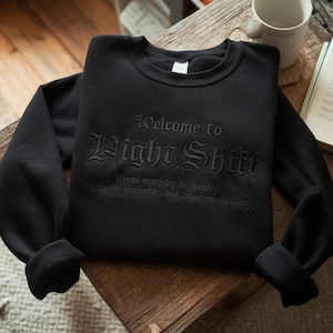 Op de afbeelding: Zwart sweatshirt met ronde hals en de tekst "Welcome to Night Shift" in gotische letters. Het sweatshirt heeft ook de tekst "Where everyday is monday, today is yesterday, and tomorrow is today."