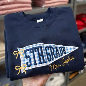 May include: A navy blue crewneck sweatshirt featuring an embroidered pennant design with a blue and white gingham pattern. The text 5TH GRADE is stitched in navy blue, with two yellow bows on the left and the name Mrs. Sophia in yellow script.